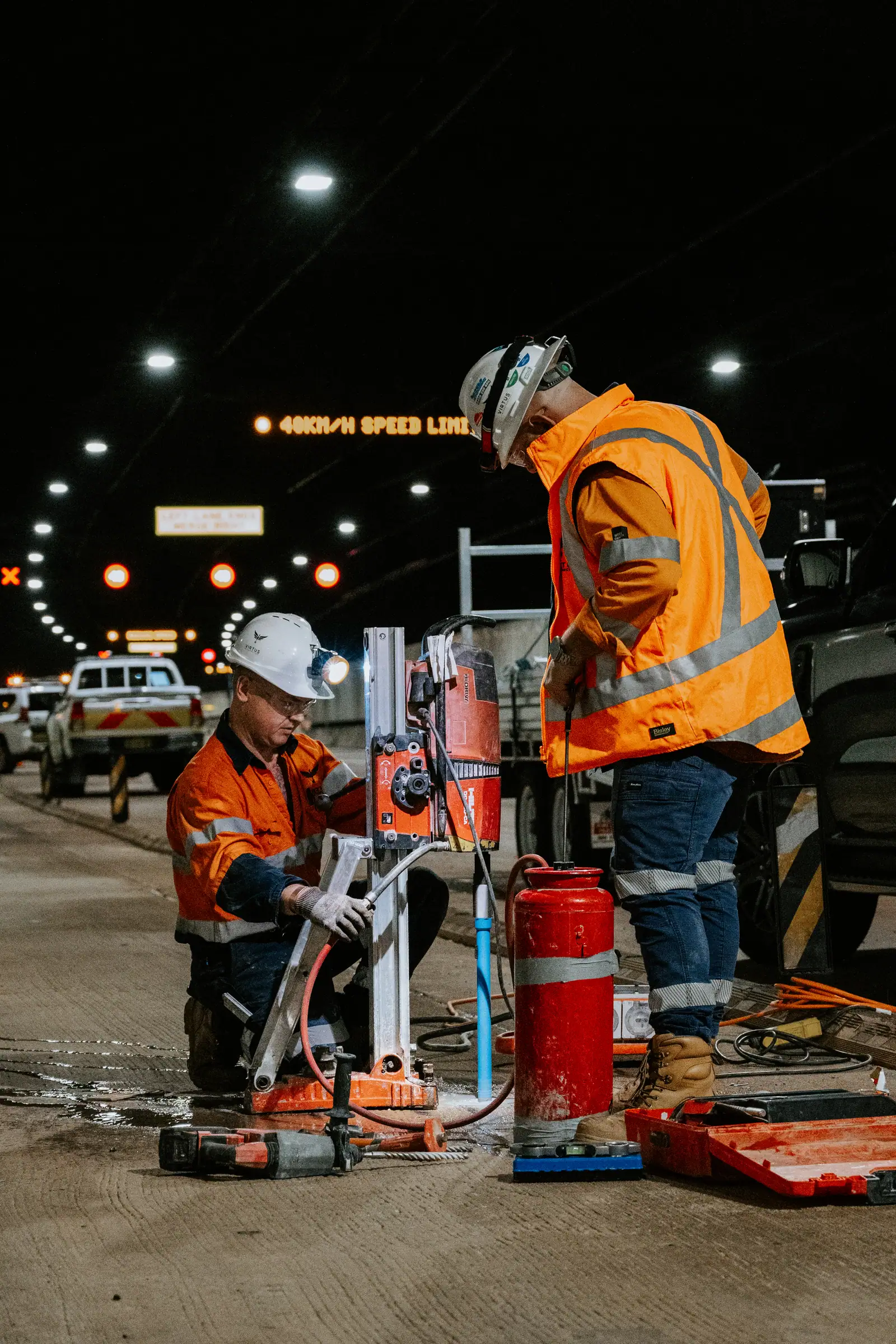 Two Virtus workers core-drilling the tunnel pavement beside a lit lane sign.